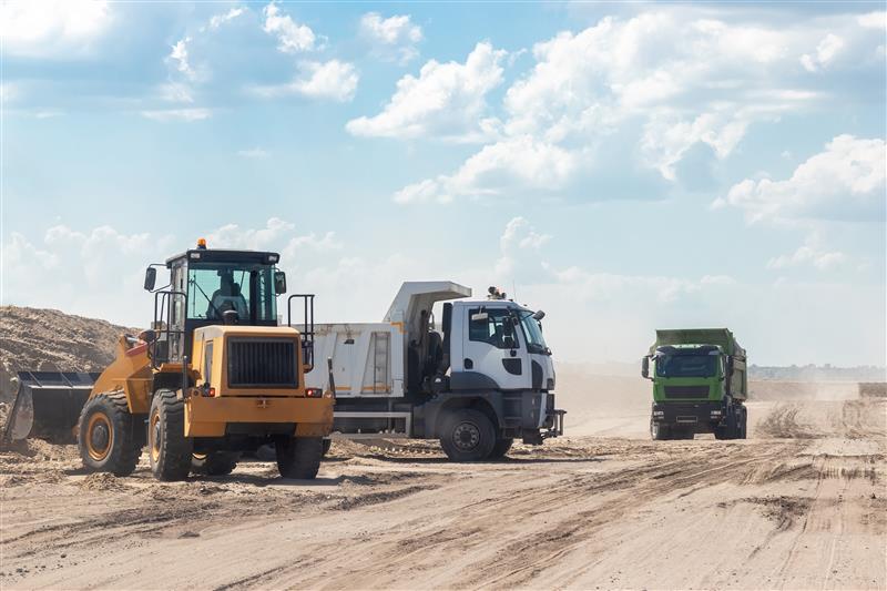 Grävmaskin och två dumprar på en sandig väg vid ett oljeraffinaderi